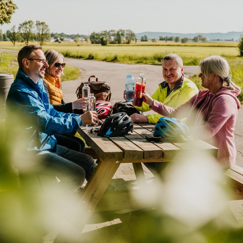 Eine Gruppe von vier Personen sitzt auf einer Bank mit Tisch und unterhält sich lachend