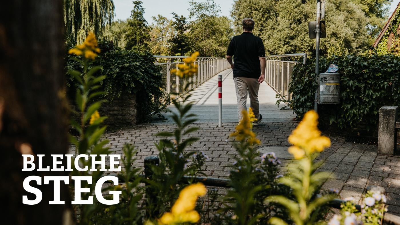Mann läuft auf dem Bleichesteg, im Vordergrund des Bildes sind blühende Blumen zu sehen 