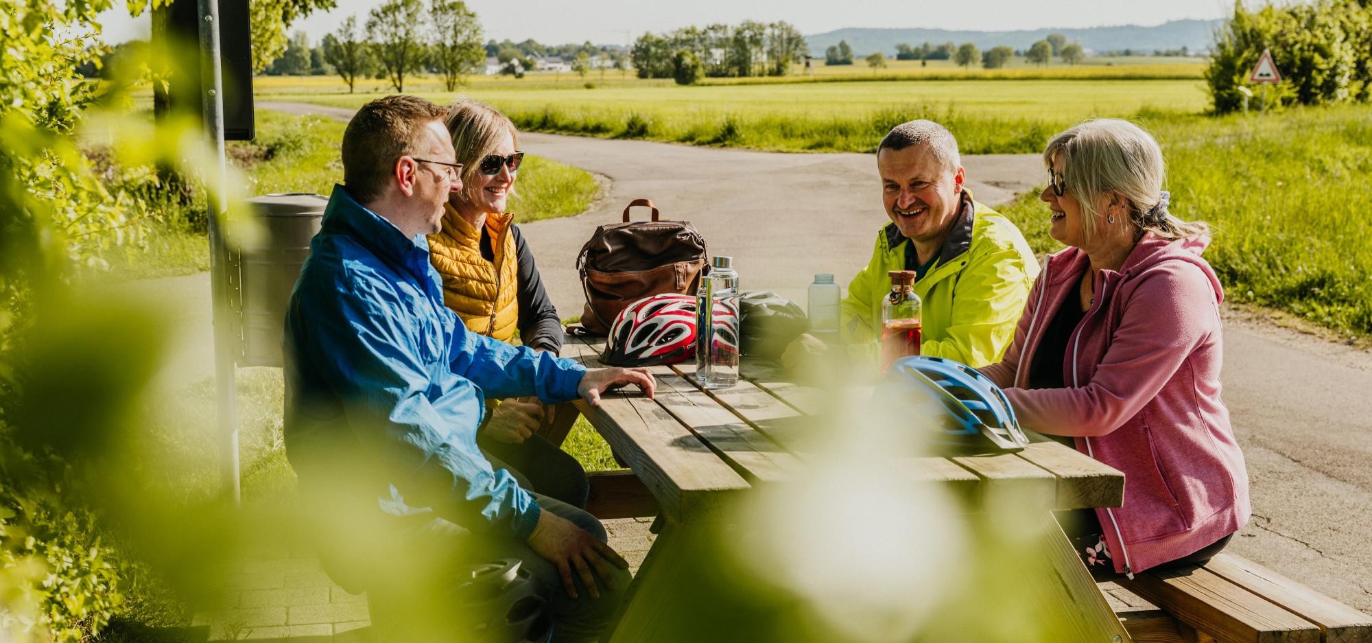 Vier Personen sitzen auf einer Sitzbank mit Tisch im Grünen und unterhalten sich lachend